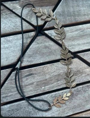 Guldigt diadem med blad - Snyggt diadem med guldfärgade blad i metall och svart elastiskt band. Perfekt för att ge din look en extra touch av stil och lite mytologisk vibe. Bladen är detaljerade och sitter längs bandet för en cool och unik känsla.