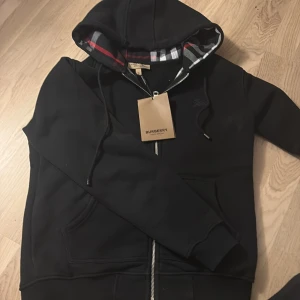 Svart hoodie från Burberry med dragkedja - Svart hoodie från Burberry i bomull med klassiskt rutigt foder i huvan, dragkedja framtill och broderad logga på bröstet. Snygg och stilren med ribbade muddar och fickor på sidorna. Perfekt för dig som vill ha en exklusiv streetstyle-look.
