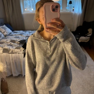 Grå ribbad half zip-tröja Vero Moda - Mysig grå ribbad tröja från Vero Moda med half zip-dragkedja och hög krage. Tröjan är stickad och har lång ärm, perfekt för lager på lager under kyliga dagar. Enkel och stilren design som passar till allt.