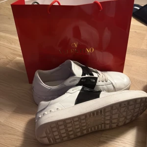 Valentino Garavani vita sneakers - Säljer ett par snygga Valentino Garavani sneakers i vitt skinn med bred svart läderdetalj över snörningen. Klassisk rund tå och platt sula, med coola nitar på sidan av sulan. Perfekt för dig som gillar exklusiva märken och stilren design.