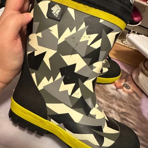 Mönstrade fodrade gummistövlar strl: 33 - Säljer ett par fodrade gummistövlar med grymt coolt grafiskt mönster i svart, grått och beige. Stövlarna har ett gult band upptill och vid sulan, samt snörning upptill för extra bra passform. Insidan är fodrad med mjukt svart foder som håller fötterna varma. Ena skosnöret saknas på skon samt står namn skrivet på lappen i skon. Några ev märken förekommer annars i fint skick.