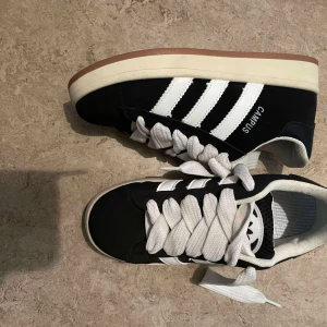 Svarta sneakers med vita ränder - Snygga svarta sneakers från Campus med klassiska vita ränder på sidorna och breda vita skosnören. Skorna har en platt sula i vitt och brunt och är perfekta för en streetwear-look. Ovandelen är i syntetmaterial och insidan är fodrad i textil.