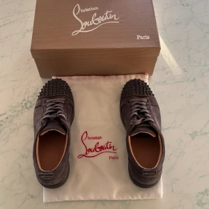 Christian Louboutin sneakers med nitar - Snygga mörkgrå sneakers från Christian Louboutin i mocka med svarta nitar på tån. Skorna har rund tå, platt sula och snörning. Insidan är i beige skinn och designen är riktigt edgy med exklusiv känsla. Perfekt för dig som vill sticka ut med lyxiga detaljer.Pris går att diskutera vid snabb affär.