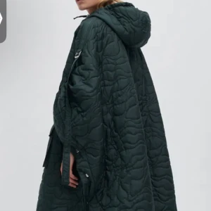 Quiltad grön pufferjacka från Ganni - Säljer en oversized quiltad pufferjacka aka poncho från Ganni i mörkgrönt. Jackan har huva, stora fickor med lock och tryckknappar, samt coola vågiga sömmar. Perfekt för dig som gillar statement-plagg och vill ha något extra till höst och vinter.