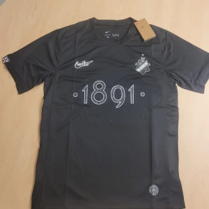 AIK Smokinglirare Edition 2025 tröja - AIK fotbollströja Smokinglirare Edition 2025 från Nike. Tröjan är mörkgrå med vita detaljer, har AIK:s klubbmärke och årtalet 1891 på bröstet. Kortärmad modell i lätt och ventilerande polyester, perfekt för match eller träning.