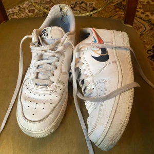 Nike Air Force 1 vita sneakers - Nike Air Force 1 sneakers i vitt skinn med klassisk rund tå och platt sula. Skorna har snörning och coola detaljer med färgglada swoosh-loggor i blått, rött och beige på sidan. Perfekt för dig som gillar streetstyle och vill ha en ikonisk sneaker i garderoben.