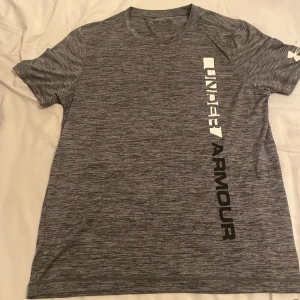 Gråmelerad Under Armour t-shirt - Snygg gråmelerad t-shirt från Under Armour med stort vertikalt logotryck i vitt och svart på framsidan. T-shirten har rund hals, korta ärmar och är tillverkad i ett lätt och funktionellt syntetmaterial som passar perfekt för träning.