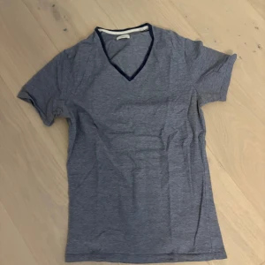 Blå v-ringad t-shirt från Bläck - Snygg blå t-shirt från Bläck med diskreta tunna ränder och v-ringad hals med mörkblå kant. T-shirten är kortärmad och i mjuk bomull, perfekt för en chill och stilren look.