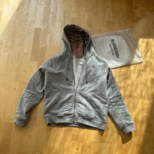 Grå hoodie från Burberry med dragkedja - Säljer en grå hoodie från Burberry med klassiskt rutigt foder i huvan och snygga detaljer med Burberry-logga på dragkedja och snörning. Tröjan har huva, snörning, två fickor fram och är långärmad. Perfekt för dig som gillar stilrena och lyxiga streetwear-plagg.