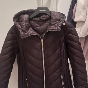 Brun pufferjacka från Michael Kors - Snygg brun pufferjacka från Michael Kors med MK-logga på ärmen och mönstrad insida med MK-tryck. Jackan har huva, guldfärgade detaljer och dragkedja framtill samt på fickorna. Perfekt för kalla dagar med sin quiltade design och stilrena look.