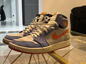 Nike Jordan 1 High i blå och orange - Nike Jordan 1 High sneakers med vit och ljusblå mocka, orange swoosh och detaljer. Klassisk hög modell med snörning och rund tå. Snygg kontrast mellan färgerna och en platt sula med orange yttersida. Perfekt för dig som gillar streetwear och unika färgkombinationer.