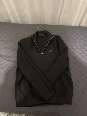 Svart half zip tröja från BOSS - Svart långärmad tröja från BOSS med half zip och ribbade detaljer. Snyggt broderad logga på bröstet och diskreta gröna inslag vid kragen. Materialet är stickat och ger en modern, clean look. Perfekt för dig som gillar stilrena plagg med premiumkänsla.