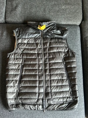 Svart quiltad dunväst Parajumpers L - Quiltad svart dunväst från Parajumpers Day Tripper med snygga detaljer som gul rem vid kragen och orange insida. Västen har två fickor med dragkedja och en markerad PJS-dragkedja framtill. Insidan är ljusgrå och materialet är lätt och syntetiskt.
