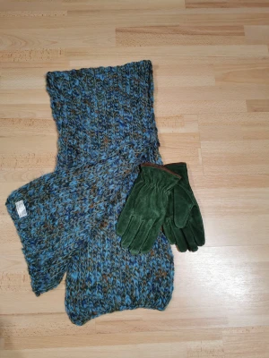  halsduk och mocka handskar  - Säljer en grovstickad halsduk i blåmelerad färg med inslag av brunt och grönt. Halsduken är tillverkad i Italien och består av 70% akryl och 30% ull. Den har ett chunky, mysigt utseende och passar perfekt för kalla dagar. Genuine Leather gröna handskar stl L 