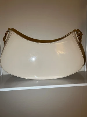 Beige axelväska från Primark med kedja - Stilren beige axelväska från Primark i glansigt syntetmaterial. Väskan har en snygg bågformad design, guldfärgade kedjedetaljer och dragkedja upptill. Perfekt storlek för det viktigaste och ger en trendig touch till din outfit. Observera lite sliten men i god skick! 