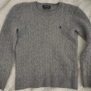 Grå stickad tröja från Polo Ralph Lauren - Kabelstickad grå tröja från Polo Ralph Lauren med rund halsringning och klassisk broderad logga på bröstet. Mjuk och skön, perfekt för lager-på-lager. Långa ärmar och ribbade muddar ger en snygg passform.