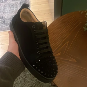Christian Louboutin svarta sneakers med nitar - Svarta sneakers från Christian Louboutin med ikonisk röd sula och coola svarta nitar på tån. Skon har snörning, rund tå och är tillverkad i skinn och mocka. Insidan är beige och sulan har Louboutin-loggan. Perfekt för dig som gillar edgy designerstil.