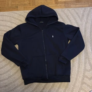 Blå hoodie från Polo Ralph Lauren - Blå hoodie från Polo Ralph Lauren med dragkedja och klassisk logga broderad på bröstet. Tröjan har huva och fickor framtill, perfekt för en avslappnad stil.