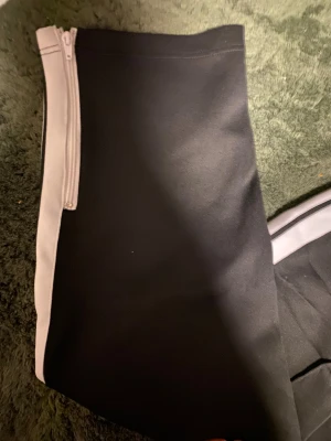 Palm Angels svarta mjukisbyxor med vita ränder - Svarta mjukisbyxor från Palm Angels med vita sidoremsor och broderad logga på vänster ben. Byxorna har elastisk midja med snörning och dragkedja vid benslut. Snygg streetwear-look med relaxed passform och sportig vibe.