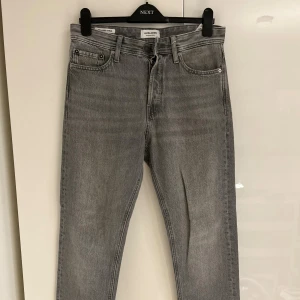 Grå jeans från Jack & Jones - Säljer ett par grå jeans från Jack & Jones, modell Relaxed Chris. De har klassisk femficksdesign, raka ben och normal passform. Jeansen är tillverkade i denim med knapp och dragkedja framtill samt detaljerade bakfickor.