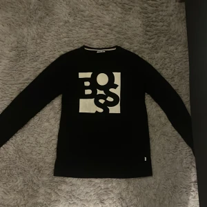 Svart långärmad BOSS tröja slim fit - Svart långärmad tröja från BOSS i slim fit med stor vit BOSS-logga tryckt på bröstet. Tröjan har en klassisk rund hals och är tillverkad i mjukt material som känns skönt mot huden. Perfekt för dig som gillar stilrena och moderna plagg.