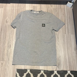 Grå t-shirt från Stone Island - Snygg grå t-shirt från Stone Island med klassisk logga på bröstet. T-shirten har rund halsringning och korta ärmar. Tillverkad i mjuk bomull som känns skön mot huden. Perfekt för en clean och stilren look. Saknar clg kod då min kompis råkade klippa av den men den är äkta