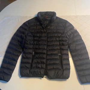 Svart Moncler dunjacka med dragkedja - Svart dunjacka från Moncler med quiltad design och två sidofickor med dragkedja. Jackan har hög krage och Moncler-logga på ärmen. Perfekt för kalla dagar och har en clean, stilren look som funkar till det mesta.