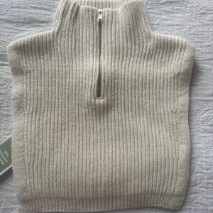 Ny. Beige ribbad stickad krage med zip - Ribbstickad beige krage med hög hals och half zip-dragkedja framtill. Perfekt att slänga över en tröja för extra värme och stil. Tillverkad i en mjuk mix av polyester, akryl och polyamid. Enkel och clean design som passar till allt.