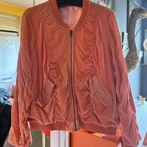 Orange bomberjacka från Odd Molly - Snygg orange bomberjacka från Odd Molly i mjuk sammet. Jackan har dragkedja framtill, två fickor med lock och coola rynkade detaljer längs ärmar och rygg. Broderad text på ena ärmen i rosa. Andvänd 1 gång.