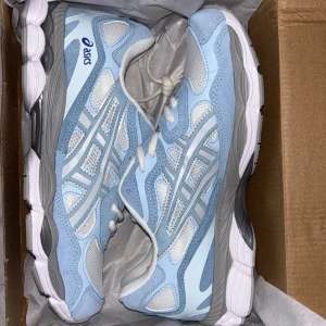 Asics sneakers i ljusblå och vitt - Snygga Asics sneakers i ljusblått och vitt med grå detaljer. Skorna har meshpaneler för bra andningsförmåga, klassisk snörning och en bekväm, stötdämpande sula. Perfekta för dig som gillar sportig stil och vill ha något fräscht till vardags.