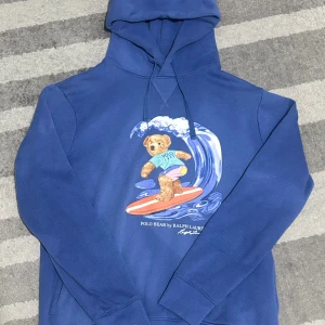 Blå Polo Bear Hoodie Ralph Lauren - Superfin blå hoodie med den ikoniska Polo Bear som surfar! 🐻🏄‍♂️ Perfekt för dig som vill sticka ut med en unik och stilren streetwear-look. Mjuk och bekväm kvalitet, klassisk Ralph Lauren-passform och en riktigt snygg print framtill.  Längd från axeln och ner: 60 cm  Bredd under armhålorna: 52 cm  📦 Skickas snabbt & välpackat.