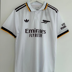 Vit Arsenal fotbollströja Adidas - Snygg vit Arsenal fotbollströja från Adidas med svarta och gula detaljer på krage och ärmslut. Tryck med Emirates Fly Better på bröstet och klubbens klassiska kanonlogga. Materialet är lätt och andas, perfekt för match eller träning.