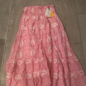 Rosa långkjol med mönster från Gina Tricot - Supersöt långkjol från Gina Tricot i rosa och vitt med blommigt mönster. Säljs inte längre! Aldrig använt! Prislapp på! Kjolen har smock i midjan och är gjord i ett lätt och luftigt bomullstyg, perfekt för varma dagar. Den har en flowy passform som ger en avslappnad och trendig look.