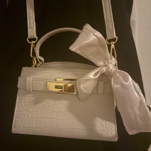 Vit axelväska med rosett och guldspänne - Säljer en stilren vit axelväska med krokopräglat mönster och snygga gulddetaljer. Väskan har både handtag och axelrem samt en stor, glansig rosett i sidenliknande material på sidan. Perfekt för dig som vill ha en trendig och elegant accessoar.