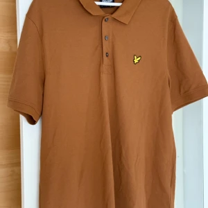 Brun pikétröja från Lyle & Scott - Säljer en brun pikétröja från Lyle & Scott med klassisk krage och tre knappar framtill. Tröjan har korta ärmar och den ikoniska gula örnloggan broderad på bröstet. Tillverkad i mjuk bomull som är skön att bära hela dagen. NYPRIS: 749kr