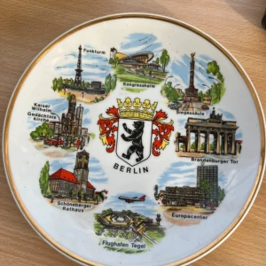 Souvenir-tallrik Berlin - Dekorativ souvenir-tallrik med motiv av Berlins kända landmärken, inklusive Brandenburger Tor och Siegessäule. Tallriken har guldkant och är i gott skick, med upphängningsanordning på baksidan. Perfekt för samlare eller som minne från Berlin.