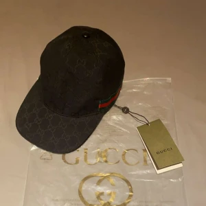 Gucci Keps  - Svart keps från Gucci med klassiskt GG-monogram över hela kepsen och ikoniska gröna och röda ränder på sidan. Justerbar rem baktill i skinn och tillverkad i Italien. Snygg och stilren accessoar för dig som gillar lyxiga detaljer.