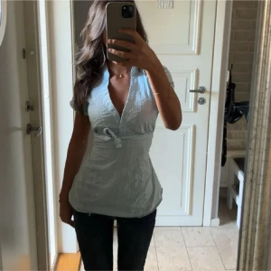 Ljusblå omlottblus med knyt - Säljer en ljusblå blus med korta ärmar och v-ringning. Toppen har knappar framtill och ett snyggt knyt i midjan som framhäver figuren. Materialet är lätt och ser ut att vara bomull, perfekt för vår och sommar.