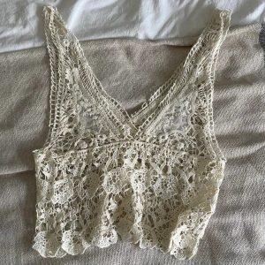 Beige spetsig croppad linnetopp - Säljer en superfin croppad linnetopp i beige med spetsdetaljer och v-ringad hals. Toppen är ärmlös och har ett vackert broderat mönster med genombruten spets som ger en boho-vibe. Perfekt för varma dagar och festivaler!