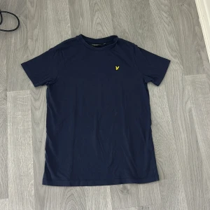 Marinblå t-shirt från Lyle & Scott - Snygg marinblå t-shirt från Lyle & Scott med klassisk rund hals och korta ärmar. På bröstet sitter den ikoniska gula fågeln broderad. Tillverkad i mjuk bomull för en skön känsla hela dagen. Fin att ha på sig, den passar för både killar och tjejer. Den är använd men fortfarande helt ren. Pris diskuteras. 