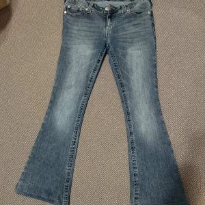 Bootcut jeans från True Religion, blå stl 31 - Nästan nya! Snygga blå bootcut jeans från True Religion med klassisk femficksdesign och ikonisk broderad logga på bakfickorna. Jeansen har låg midja, slitningar och kontrastsömmar som ger en cool vintagekänsla. Perfekt för dig som gillar moderna jeans. De är uppsydda med tråd som är lätt att sprätta bort. Jag är 1,64 m så passar någon upp till ca 1,70 minst.  