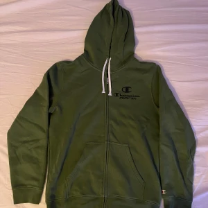 Grön hoodie med dragkedja från Champion - Grön hoodie från Champion med hel dragkedja, vit snörning i huvan och broderad logga på bröstet. Mjuk bomullsblandning, känguruficka framtill och klassisk Champion-detalj på ärmen. Perfekt för chill eller träning.