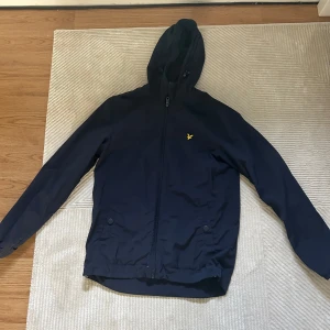 Mörkblå vindjacka från Lyle & Scott - Snygg mörkblå vindjacka från Lyle & Scott med huva och dragkedja framtill. Jackan har två fickor med knappar och den klassiska gula loggan på bröstet. Perfekt för blåsiga dagar och enkel att matcha med det mesta.