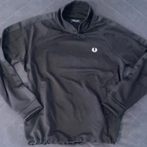 Svart Fred Perry half zip tröja - Svart långärmad tröja från Fred Perry med half zip och hög krage. Tröjan har en broderad logga på bröstet och ribbade muddar. Materialet känns mjukt och sportigt. Dragsko nertill för justerbar passform. Storlek XL men passar mer L
