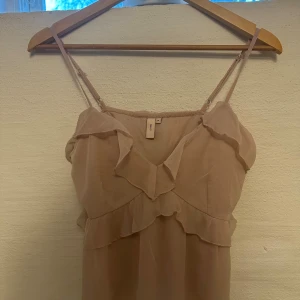 Beige volangklänning från Nelly - Supersöt beige klänning från Nelly med tunna axelband och volangdetaljer både fram och bak. Lätt och luftig modell i ett skirt material som ger en romantisk vibe. Perfekt för varma dagar när du vill känna dig extra fin.