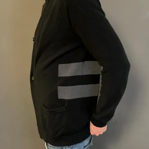 Svart cardigan från Emilio - Svart kofta med gråa horisontella ränder på sidorna. Tröjan har ribbad krage och muddar, knappar framtill och två fickor. Materialet är stickat och koftan har en normal passform med långärmade ärmar. Perfekt för dig som gillar stilrena detaljer.