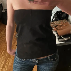 Svart bandeau tubtopp - Säljer en svart bandeau tubtopp med stilren och enkel design. Toppen är axelbandslös och har en rak, tight passform framtill och spetsig nederkant i sidorna. Perfekt att styla med jeans eller kjol för en trendig look.