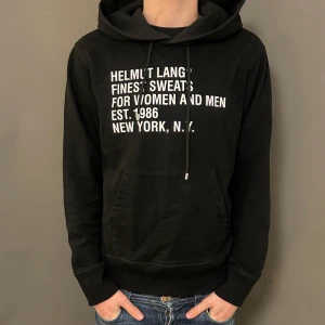 Svart hoodie från Helmut Lang - Svart hoodie från Helmut Lang med vit textprint på bröstet. Tröjan har huva med snörning, stor magficka och ribbade muddar. Materialet är mjuk bomull och passformen är normal. Perfekt för dig som gillar streetwear och enkel stil.