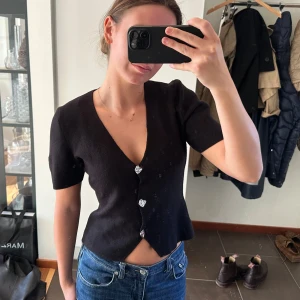 Svart stickad topp från Gina Tricot - Svart stickad topp med kort ärm och v-ringning från Gina Tricot. Toppen har tre dekorativa knappar framtill och en lätt croppad passform. 
