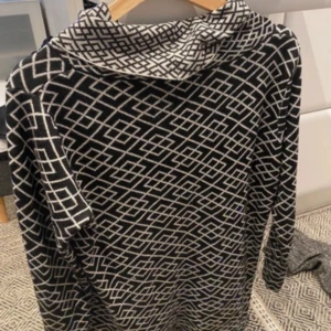 Svartvit polotröja med grafiskt mönster - Svart polotröja med vitt grafiskt rutmönster över hela tröjan. Tröjan har hög krage och långa ärmar, perfekt för dig som gillar en stilren men ändå unik look. Materialet känns mjukt och passar bra till kyligare dagar.